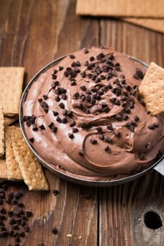 a bowl filled with chocolate frosting surrounded by crackers and grahams on a wooden table