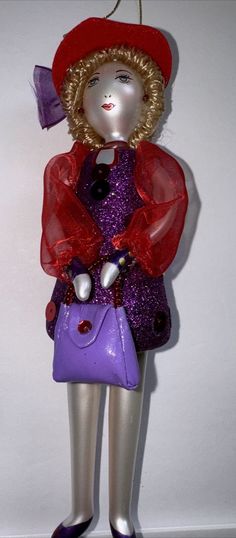 a doll is wearing a purple dress and red hat, holding a purple handbag