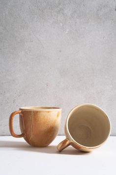 two coffee mugs sitting next to each other on a white counter top, one is brown and the other is beige