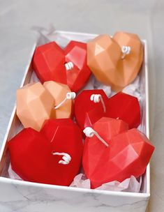 four heart shaped candles in a white box on a marble counter top, one is red and the other is orange