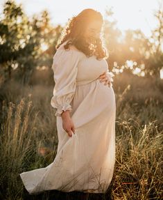 a pregnant woman standing in tall grass with her hands on her hips, wearing a white dress