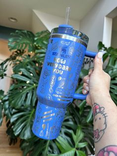 a person holding up a blue tumbler cup with writing on it and a plant in the background