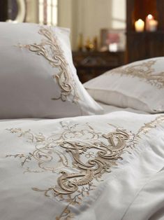 a bed with white sheets and gold embroidered designs on the pillow cases, along with candles