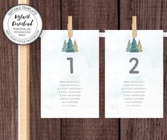 two wedding seating cards with pine trees on them, hanging from clothes pins in front of a wooden background