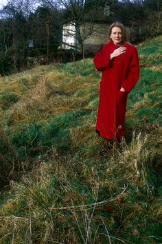 a woman in a red coat is standing on a grassy hill with her hands together