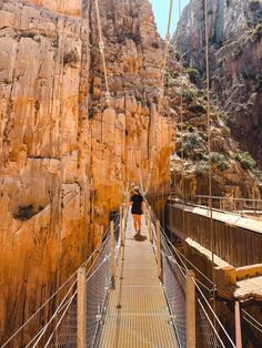 a person walking across a suspension bridge in the mountains with cliffs behind them on a sunny day