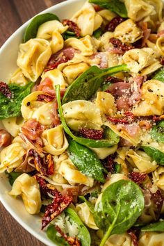 a white bowl filled with pasta, spinach and other vegetables on top of a wooden table