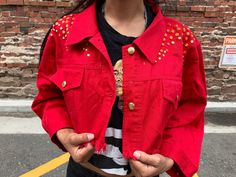 a woman wearing a red jacket with gold studs on the sleeves and shoulders, standing in front of a brick wall