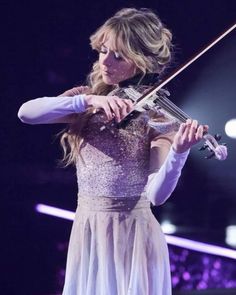 a woman in a white dress holding a violin on top of a stage with lights behind her