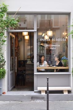 two women sitting at a table in front of a store window with plants growing on the outside