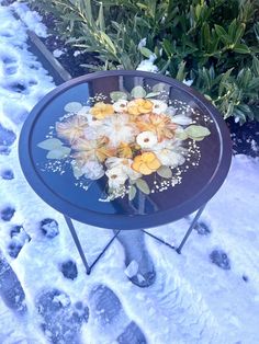 a glass table with flowers painted on it in front of snow covered ground and shrubbery