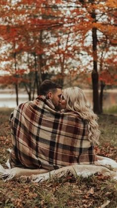 a man and woman wrapped up in a blanket sitting on the ground with trees behind them