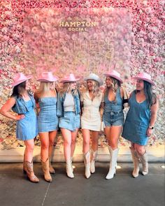 four women in denim dresses and cowboy hats posing for a photo with a floral wall behind them