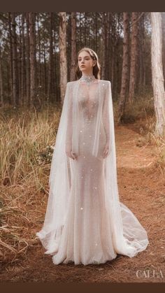 a woman in a white dress standing in the woods wearing a long cape and veil