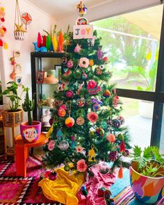 a brightly colored christmas tree with ornaments on it in front of a window and potted plants