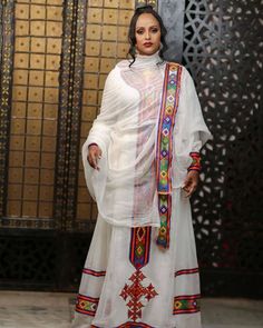 a woman standing in front of a wall wearing a white dress with colorful trims