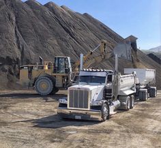 two dump trucks are parked in front of a large rock formation with mountains in the background