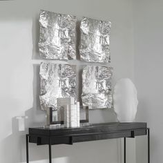 a black table topped with a white vase next to two silver wall art pieces on top of it