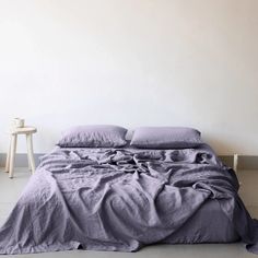 an unmade bed with purple linens in a white room next to a table