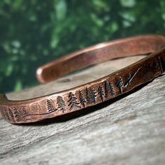 A rustic handmade bracelet that celebrates the beauty of nature and the majesty of the forest with the mountains and trees hand stamped around the rustic copper cuff.   Perfect for the mountain climber or hiker in your life.  This engraved copper bracelet is hand forged and finished with a hammered finish to give it a rustic look, perfect for men and women alike.  Each bracelet is handmade, and no two will be exactly alike. Also please be aware that colors may vary due to viewing screens. If you Mens Copper Bracelet Western, Copper Bangles Handmade, Stamped Cuff Bracelets, Engraving Ideas Jewelry, Copper Smithing, Hammered Copper Jewelry, Hammered Metal Jewelry, Mountain Bracelet, Mens Copper Bracelet