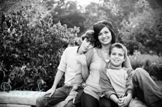 a woman and two boys sitting on a bench in front of some bushes with their arms around each other