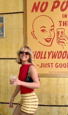 a woman in a short skirt holding a drink and posing for the camera with a no poop sign behind her