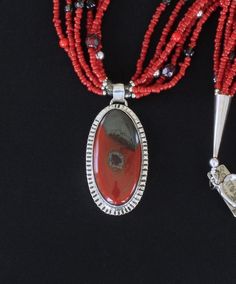 This appealing Necklace showcases a Rainbow Brecciated Jasper and Sterling Silver Pendant by Nevada Silversmith Jim Daggett. The Oval Pendant Stone is a vibrant brick red with black markings. It is wrapped in a raised Sterling Silver Bezel and set on Sterling Sheet cut to mirror the shape of the Stone. The Sheet has linear stamping that is radiant in reflected light. The Pendant drapes from a small Sterling Silver Cylinder Bail and is stamped on the back with the Artist's hallmark and "Sterling. Red Beaded Pendant Necklace With Natural Stones, Red Gemstone Beads And Cabochons For Gifts, Red Hand-strung Beads For Jewelry Making, Handmade Artisan Red Beads, Gems, And Cabochons, Red Natural Stones Beads And Cabochons For Gifts, Artisan Red Necklaces With Polished Beads, Artisan Red Necklace With Polished Beads, Artisan Red Gemstone Beads, Red Oval Gemstone Bead Jewelry