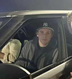 a man sitting in the passenger seat of a car next to another man wearing a new york yankees hat