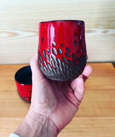 a hand holding a red and black cup on top of a wooden table next to a bowl