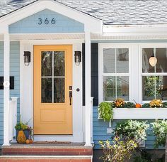 a blue house with white trim and yellow front door has pumpkins in the window boxes