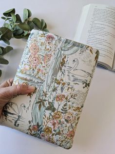 a person holding an open book on top of a table next to flowers and leaves