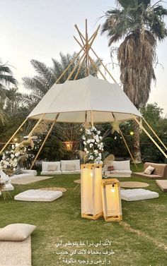 a white tent with pillows and lights on it in the middle of a lawn surrounded by palm trees