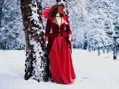 a woman in a long red dress standing next to a tree with snow on it