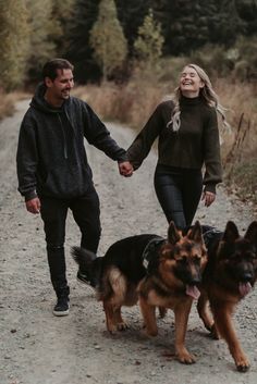 a man and woman holding hands walking with two dogs on a dirt road in the woods