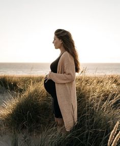 a pregnant woman standing in tall grass on the beach at sunset with her belly exposed