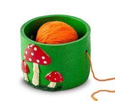 a ball of yarn sits in a green cup with mushrooms on the side and orange yarn next to it