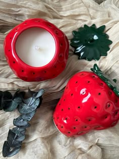 a red strawberry shaped candle sitting on top of a white blanket next to a green plant