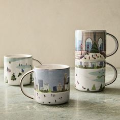 three coffee mugs sitting on top of a counter next to each other with different scenes painted on them