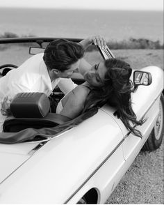 a man and woman laying on top of a convertible car near the ocean in black and white