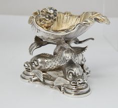 an ornate silver bowl with fish and flowers on it's base, sitting on a white surface