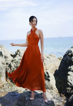 a woman in an orange dress standing on rocks near the ocean with her arms outstretched