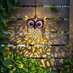 an owl lamp hanging from the side of a wooden fence with green leaves surrounding it