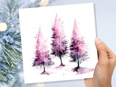 a hand holding up a card with watercolor trees on it in front of a christmas tree