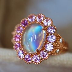 "Make a statement with this one-of-a-kind Edwardian-inspired rainbow moonstone ring. The center stone is a radiant rainbow moonstone, surrounded by a halo of pink and purple sapphires. The 14K rose gold band features a delicate leaf design on the sides, adding to the vintage elegance of the piece. Perfect for special occasions or everyday wear, this ring is sure to become a treasured addition to any jewelry collection The size is in US 7 made of solid 14K rose gold can be sized up or down for $6 Pink Moonstone Engagement Ring, Moonstone Multi-stone Opal Ring As Gift, Multi-stone Moonstone Opal Ring For Gifts, Pink Opal Gemstone Ring For Anniversary, Pink Opal Ring With Accent Stones As Gift, Unique Moonstone Gemstone Ring For Anniversary, Fine Jewelry Rose Gold Moonstone Ring, Unique Moonstone Ring With Gemstone Accents For Anniversary, Unique Moonstone Ring With Accent Stones