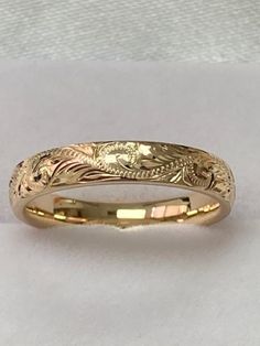 a yellow gold wedding band with an intricate design on the front and side, sitting on a white surface