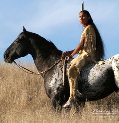 a native american woman sitting on top of a black and white horse in a field