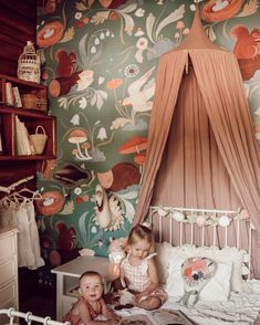 two small children sitting on a bed in a room with floral wallpaper and curtains