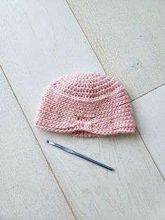 a pink knitted hat laying on top of a wooden floor next to a knitting needle