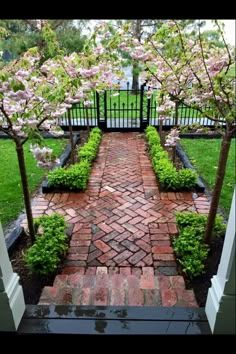 a brick pathway with trees and flowers in the background, leading to a gated area