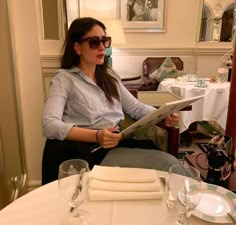 a woman sitting at a table in a restaurant reading a paper with glasses on it